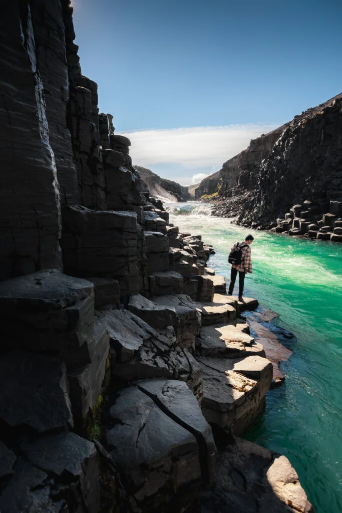 A hiker on the rocky bank below the basalt columns of Stuðlagil Canyon