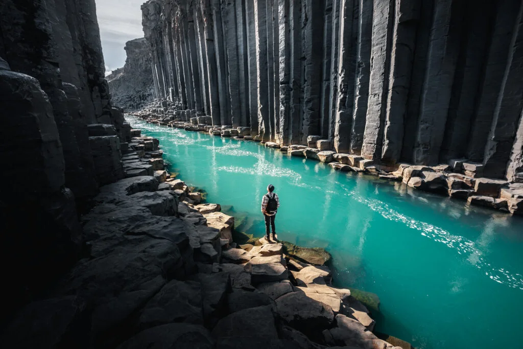 A hiker beside turquoise water and tall basalt columns in Stuðlagil Canyon